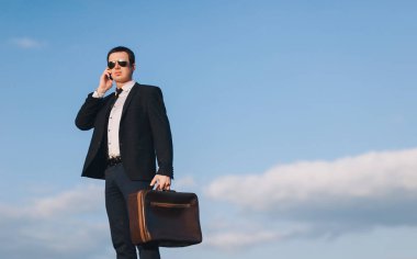 Takım elbiseli, kravat ve güneş gözlüklü yakışıklı genç işadamı mavi gökyüzü arka planında duruyor ve cep telefonuyla konuşuyor. Boşluğu kopyala.