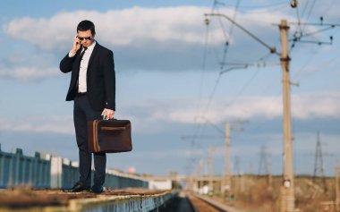 Takım elbiseli, kravat ve güneş gözlüklü yakışıklı bir iş adamı bir tren istasyonunda duruyor ve cep telefonuyla konuşuyor. Bulutlu akşam masmavi gökyüzü.