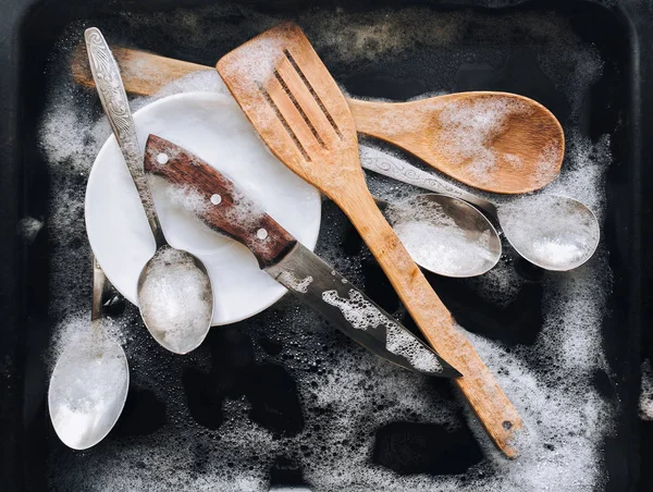 Bir tabak, bir bıçak, ahşap mutfak spatulaları ve siyah bir fırın tepsisindeki deterjan köpüğünde kaşıklar. Bulaşık yıkama kavramı. Üst görünüm.