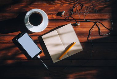 Beyaz ekranlı siyah akıllı telefon, kağıt defter, kalem, kulaklık ve kahve kahverengi ahşap arka plan maununda güneş ışığı lekeleri olan bir kahve. Uzayı kopyala, model. Müzik ve yazım konsepti.