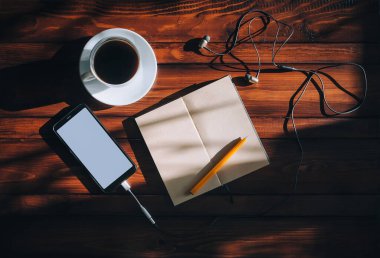 Black smartphone with white screen, paper notebook, pen, headphone and cup of coffee lies on a brown wooden background mahogany with spots of sunlight. Copy space, mockup. Music and writter concept. clipart