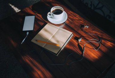 Kağıt defter, kalem, beyaz ekranlı siyah akıllı telefon, kulaklık ve kahve kahverengi ahşap arka plan maununda güneş ışığı lekeleri olan bir fincan kahve. Uzayı kopyala, model. Müzik ve yazım konsepti.