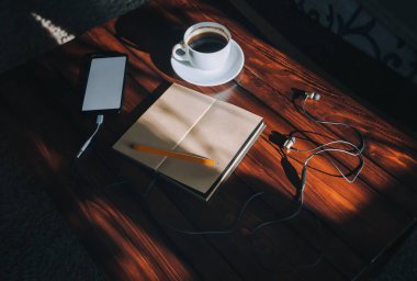 Paper notebook, pen, black smartphone with white screen, headphone and cup of coffee lies on a brown wooden background mahogany with spots of sunlight. Copy space, mockup. Music and writter concept. clipart
