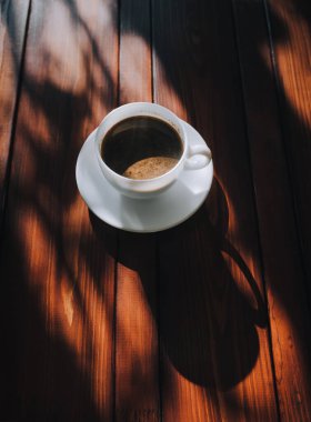 Kahveli ve çay tabaklı beyaz bir kupa, koyu kahverengi bir ahşap maun masanın üzerinde, güneşin ışığında duruyor ve koyu bir gölge bırakıyor. Yalnız adam sabah kahve konsepti.