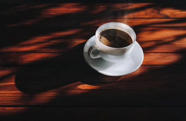 コーヒーとソーサー付きの白いカップは 太陽の光の下で濃い茶色の木製のマホガニーテーブルの上に立ち 暗い影を投げました ロンリーマンモーニングコーヒーのコンセプト — ストック写真