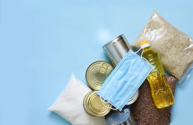 Yemek servisi. Mavi arka planda gıda bağışı ve maske: yağ, karabuğday, aris, şeker, konserve yiyecek. Evinde otur. Karantina.
