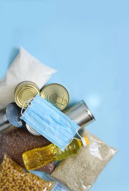 Yemek servisi. Mavi arka planda gıda bağışı ve maske: yağ, karabuğday, aris, şeker, konserve yiyecek. Evinde otur. Karantina.