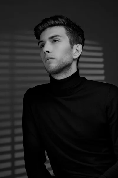 Stylish young man in a black sweater sits near blinds and thinki