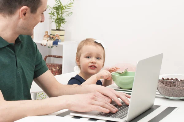 爸爸在家照顾一个小女儿 他给了她牛奶麦片 并开始使用笔记本电脑工作 生活方式 男子在家庭中的现代作用 — 图库照片