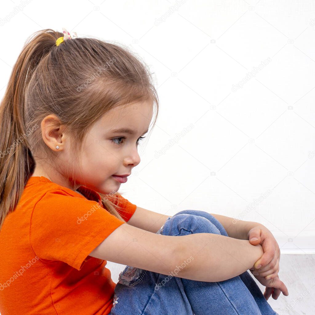 Shot of a  little smiling girl wearing orange shirt and jeans sitting on the floor clasping her knees with her hands against white background with copy space in studio