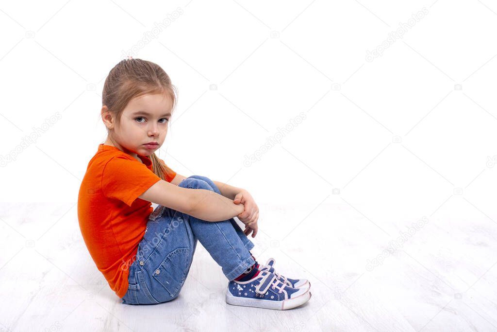 Shot of a  little upset girl wearing orange shirt and jeans sitting on the floor clasping her knees with her hands against white background with copy space in studio