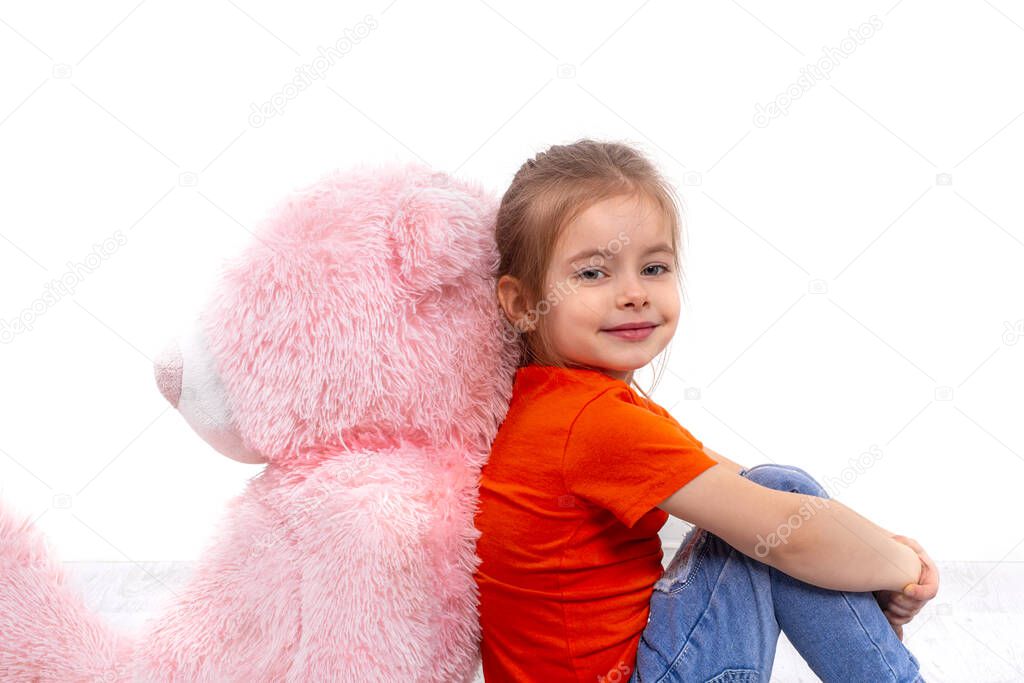 Shot of a  little smiling girl wearing orange shirt and jeans sitting wih big pink toy bear on the floor clasping her knees with her hands against white background  in studio
