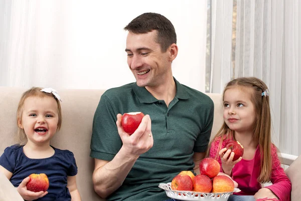 Shot of  young dad with two little daughter spending time at home.  They have fun eating apples. The concept of family values.  Life style.  The modern role of men in the family.