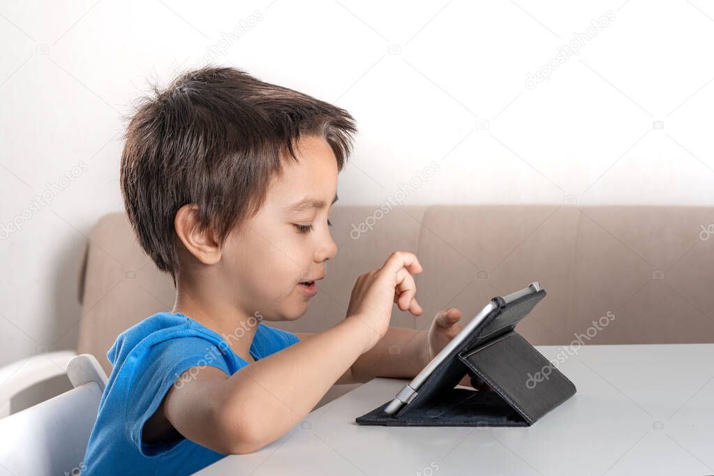 Shot of little fun boy sitting at a table at home and browsing the internrt on a  tablet. Modern lifestyle.  