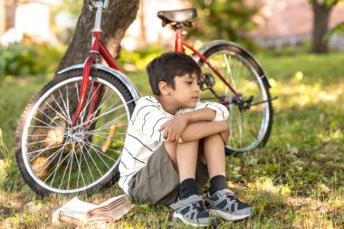 Bahçedeki çimlerin üzerinde oturan ve bir şeye üzülen genç bir çocuğun fotoğrafı, bir ağacın yanında duran kırmızı bir bisiklet, çimlerin üzerinde yatan bir kitap. Mutlu çocukluk konsepti, yaz tatili