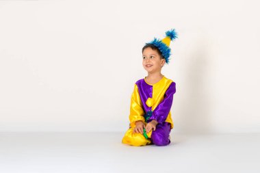 Stüdyoda, atölyede beyaz arka planda oturan palyaço kostümü giymiş küçük bir çocuğun karnaval fotoğrafı.. 