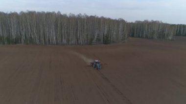 Bir traktör ormanın arka planında büyük bir tarlayı sürüyor. Bahar. Yapraksız ağaçlar. İyi akşamlar.