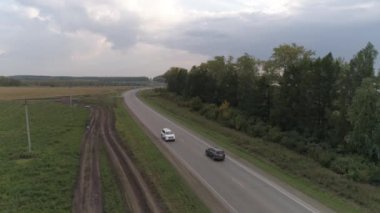 Yoldaki beyaz düğün arabalarının hava manzarası. Yaz, akşam. Yolun bir tarafında bir tarla var, diğer ağaçlarda.