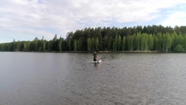 空中的湖景环绕着一片森林 两名渔民乘坐着一艘橡胶充气船驶向岸边 一个渔夫划船 另一个站着 码头上还有其他拿着钓竿的渔民 阳光灿烂的夏日 — 图库视频影像