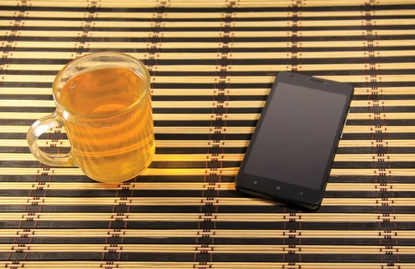 Smartphone nero e tazza di tè su un tappetino di bambù — Foto Stock