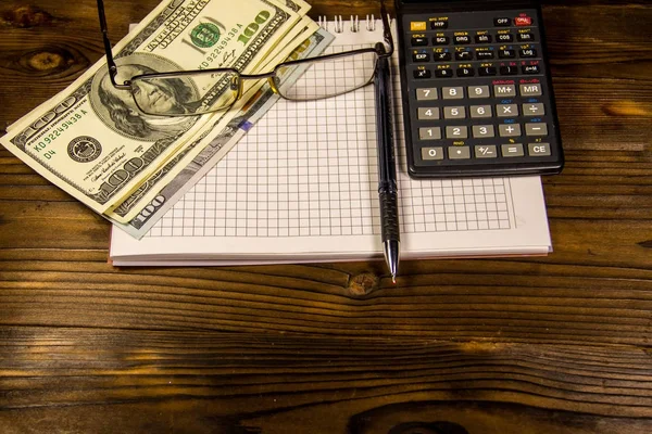 Notitieboekje met dollars, pen, bril en Rekenmachine op houten bureau — Stockfoto
