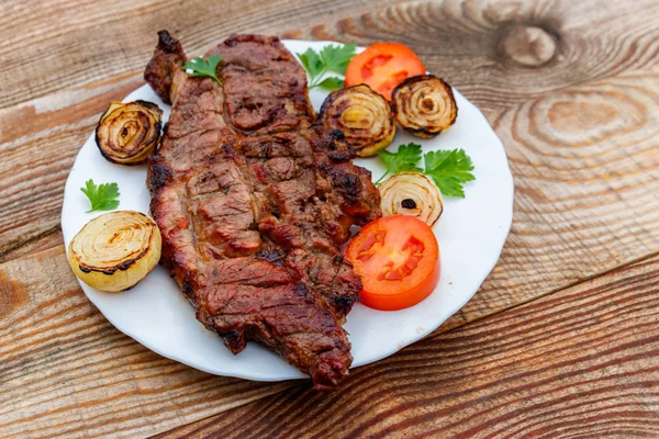 Mięso wieprzowe z grilla z cebulą, pomidorami i pietruszka w biały talerz na drewnianym stole — Zdjęcie stockowe