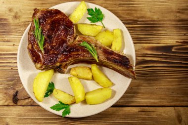 Kemiğin üstünde biftek, fırında patates ve ahşap masada otlar.