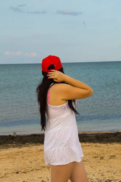 白いドレスと海のビーチに立つ赤いキャップの美しいブルネットの女の子。夏休みのコンセプト — ストック写真