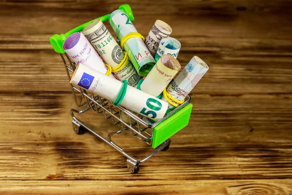 Carrito de compras con dinero de diferentes países: euros, dólares, hryvnia ucraniana, rublos rusos, libras egipcias sobre fondo de madera —  Fotos de Stock