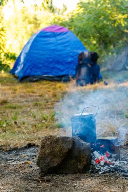 Kamp ateşinde yemek pişirmek. Arka planda çadır ve sırt çantası