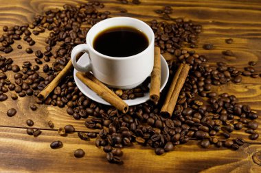 Bir fincan kahve, kavrulmuş kahve çekirdekleri ve ahşap masada tarçın çubukları.