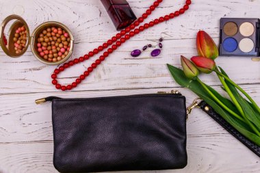 Beyaz ahşap arka planda kadın aksesuarları ve makyaj malzemeleri. Debriyaj çantası, parfüm, göz farı, ruj topları, kolye, küpeler ve laleler. Güzellik ve moda kompozisyonu. Üst manzara, düz uzanma