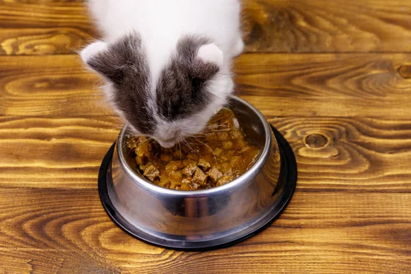 Маленький кошеня їсть свою їжу з металевої чаші на дерев'яній підлозі — стокове фото