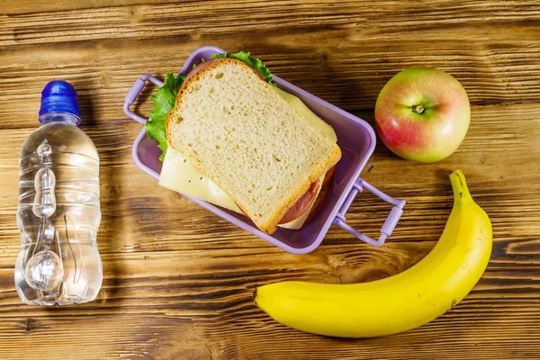 Scatola con panini, bottiglia d'acqua, banana e mela su un tavolo di legno. Vista dall'alto — Foto Stock