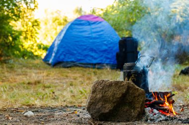 Kamp ateşinde yemek pişirmek. Arka planda çadır ve sırt çantası