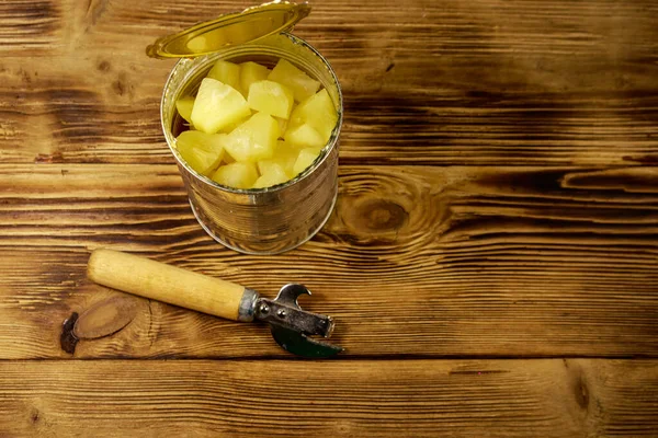 Opened tin can of pineapple pieces and can opener on wooden table