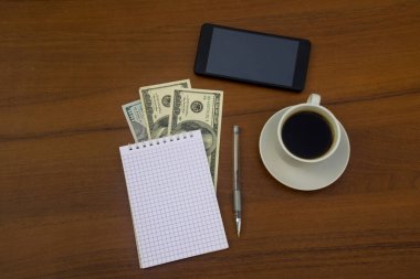 Bir fincan kahve, akıllı telefon, dolar, not defteri ve tahta masadaki kalem. Mali planlama kavramı