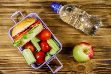Bir şişe su, elma ve beslenme çantası, içinde hamburgerler ve ahşap bir masada taze sebzeler. Üst görünüm