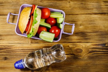 Bir şişe su ve hamburgerli beslenme çantası ve ahşap bir masada taze sebzeler. Üst görünüm