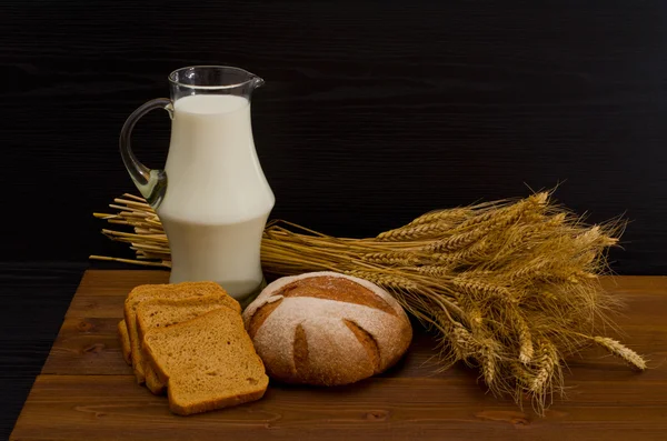 Кувшин молока, круглый ржаной хлеб, сноп пшеницы, ломтики хлеба на деревянном столе — стоковое фото
