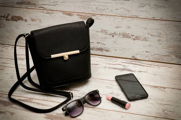 Kleine schwarze Damenhandtasche, Sonnenbrille, Handy und Lippenstift auf Holzgrund. Modekonzept — Stockfoto