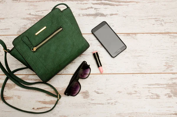 Green ladies handbag, sunglasses, phone and lipstick on wooden background. Fashionable concept, top view