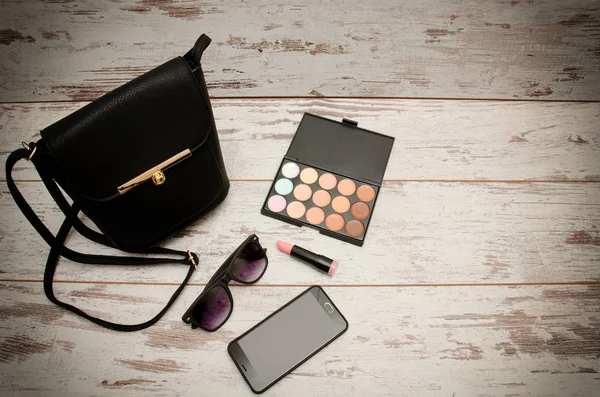 Little black ladies handbag, eye shadow, sunglasses, phone and lipstick on wooden background. Fashion concept. Top view, space for text
