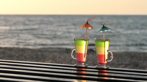 Two cocktails with straws on a wooden tabletop into the sunset on the beach, the sea in the background — Stock Video