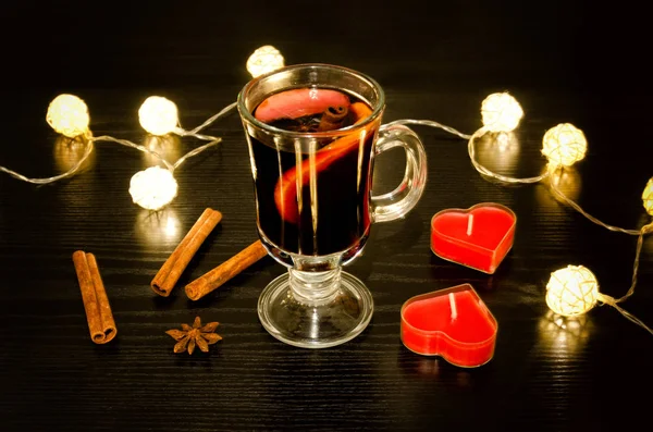 Taza de vino caliente con especias, velas en forma de corazón, palitos de canela, anís estrellado. Iluminación de faroles de ratán sobre una mesa de madera negra — Foto de Stock