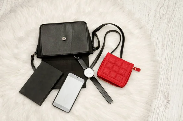 Offene schwarze Tasche mit abgeworfenen Sachen, Notizbuch, Handy, Uhr und rotem Portemonnaie. das weiße Fell auf dem Hintergrund, Draufsicht. Modekonzept — Stockfoto