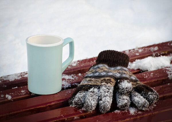Blue empty mug and women's knitted gloves on a bench in the winter, snow in the background. Close-up