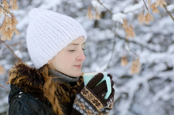 Chica en un sombrero blanco con una taza de té entre las ramas de los árboles cubiertos de nieve, vista lateral —  Fotos de Stock