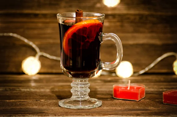 Caneca de vinho quente com especiarias de perto. Vela na forma de um coração em uma mesa de madeira, lanternas no fundo — Fotografia de Stock