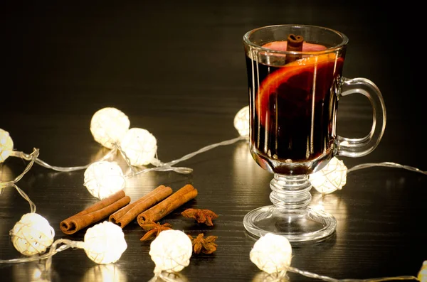 Mug of mulled wine with spices, cinnamon sticks, star anise. Illumination of rattan lanterns on a black wooden table — Stock Photo, Image